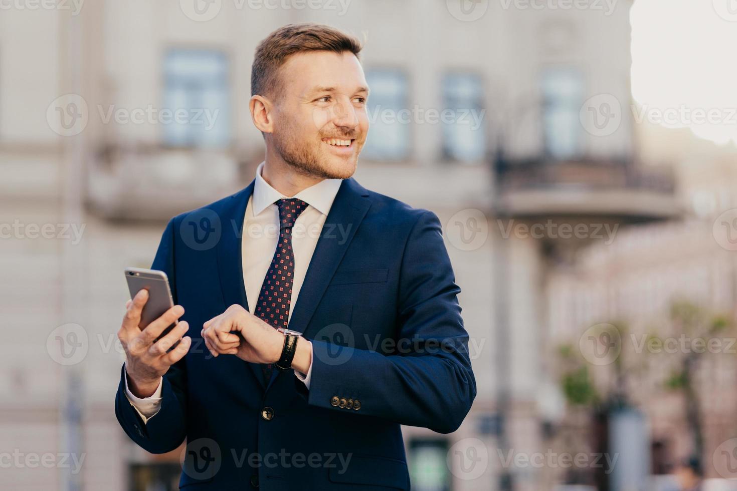 Horizontal shot of successful unshaven male CEO looks for interesting multimedia files on smart phone, downloads or updates new programs, stands against blurred city background, looks happily aside photo