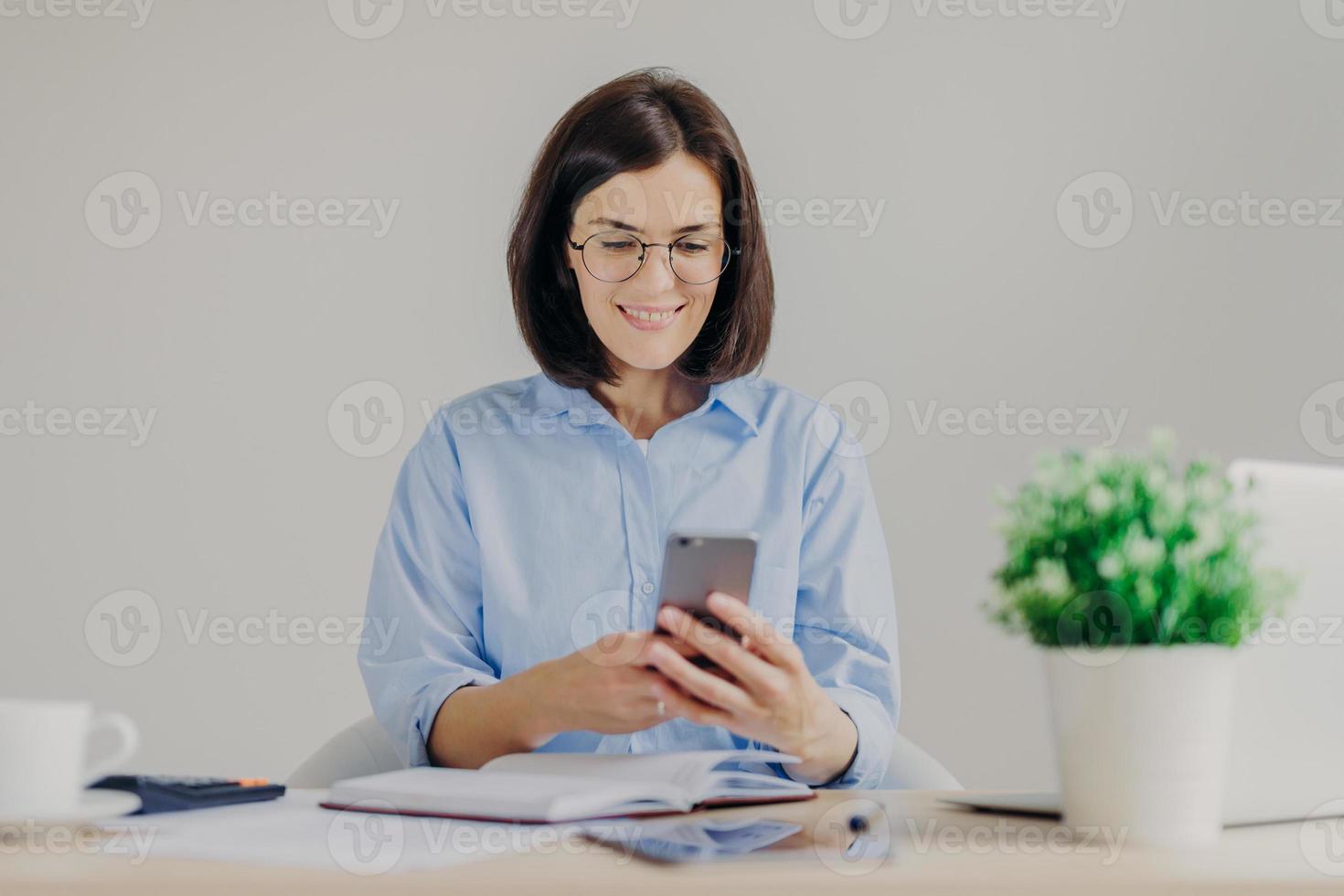 Happy female employee works on developing new project in office, searches information on websites via modern electronic gadgets, uses free internet connection in her cabinet, has happy expression photo