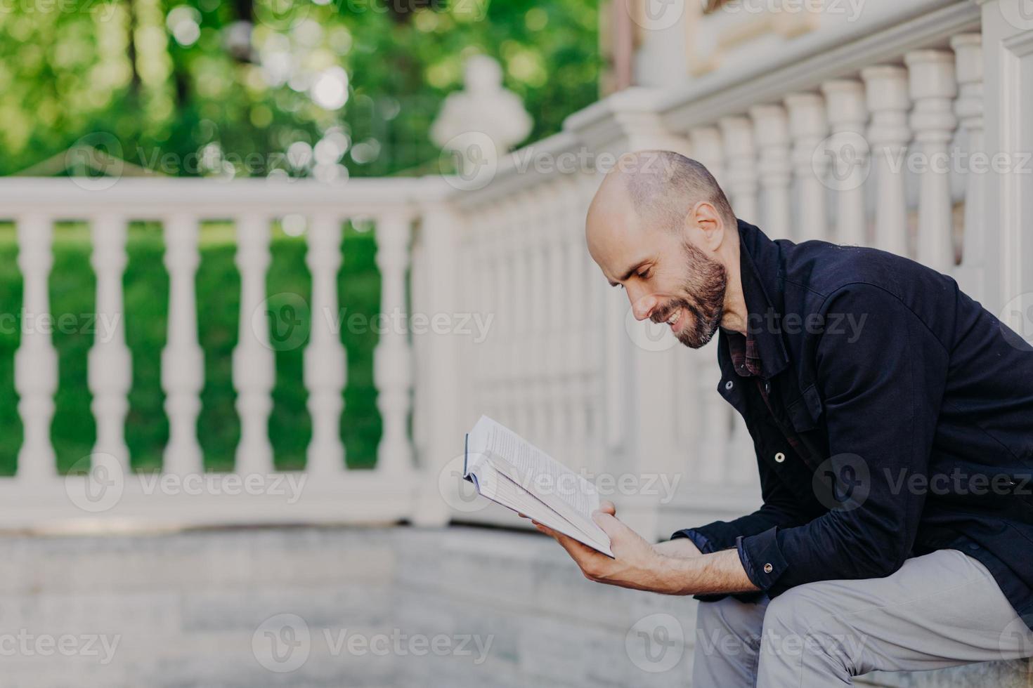 la foto de un hombre barbudo calvo de mediana edad se inclina de cerca para reservar, tiene una expresión positiva, se sienta contra un balcón blanco, tiene un rastrojo oscuro, está de buen humor y le gusta leer. el hombre lee una historia romántica