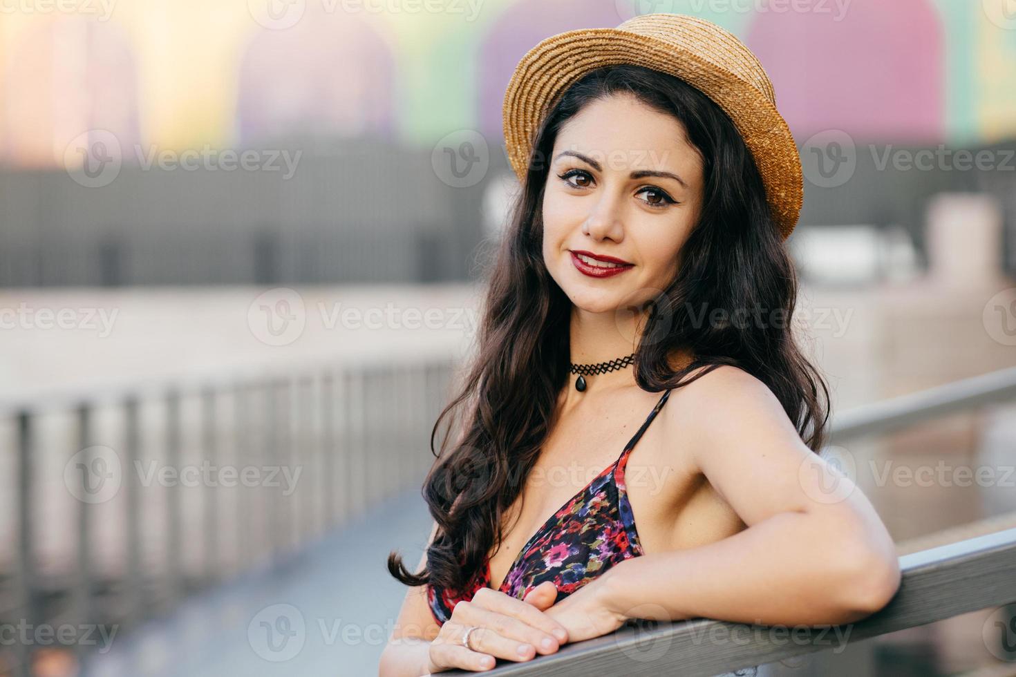 mujer encantadora con cabello largo oscuro, apariencia atractiva con sombrero de verano y vestido de excursión en la gran ciudad posando en el puente. joven viajera caminando por la calle con una mirada encantadora foto