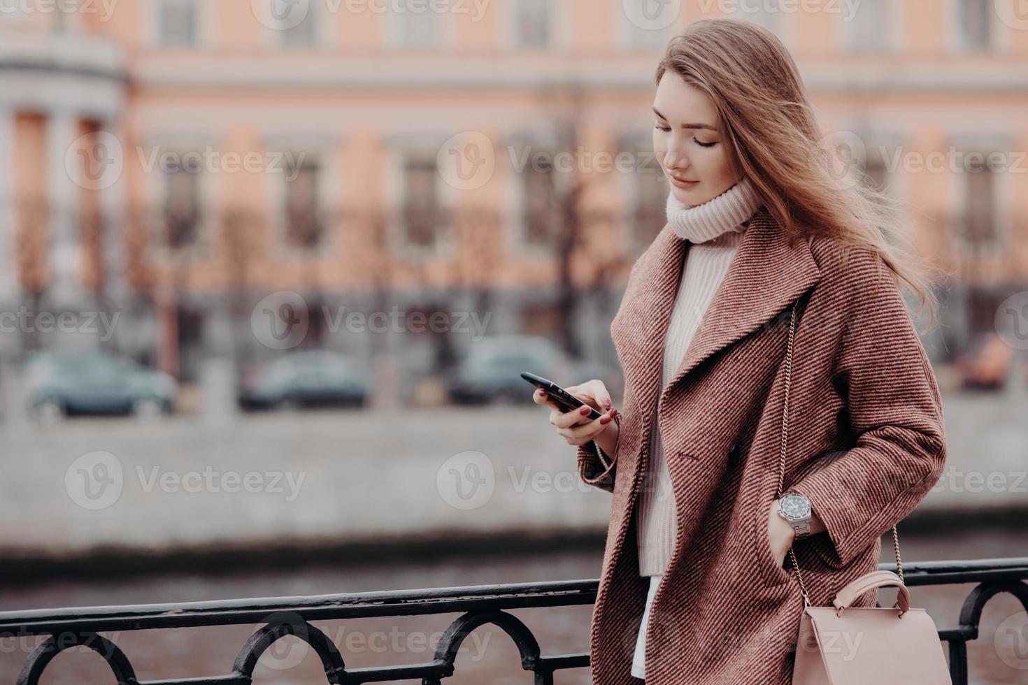Beautiful woman in warm coat, holds modern cell phone, messages in social networks, connected to free wifi, walk in city, street style, blurred background with copy space for your advertisement photo