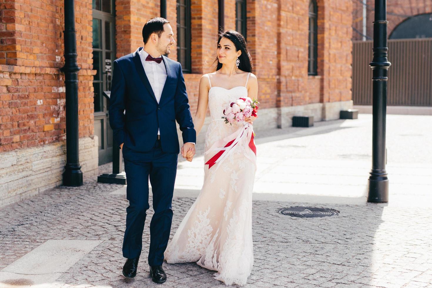 People, relationship and love concept. Brunette attractive bride in beautiful white dress, holds bouquet, keeps her lovers hand, have pleasant conversation, enjoy togetherness and calmleness photo