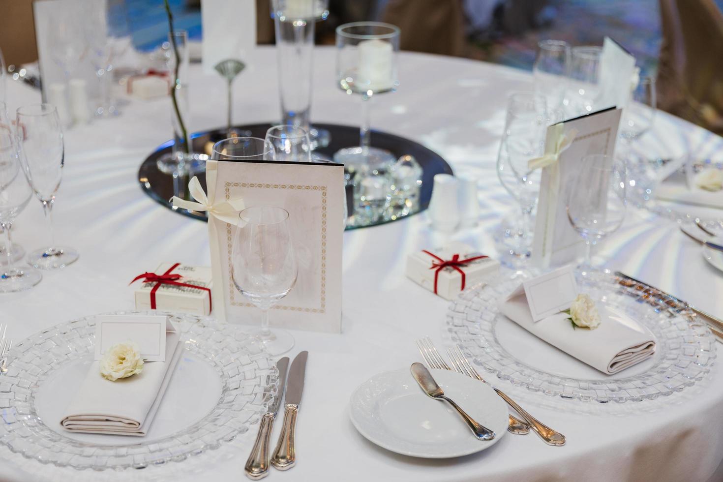 mesa redonda servida con platos vacíos, cuchillos, tenedores, tarjetas y servilletas, cubierta con manteles blancos. cubiertos, cristalería en mesa festiva. foto