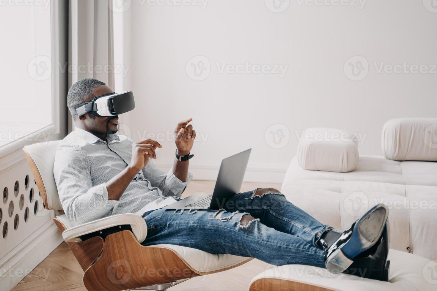 African businessman in VR glasses interact with virtual reality sit in armchair at laptop. High tech photo