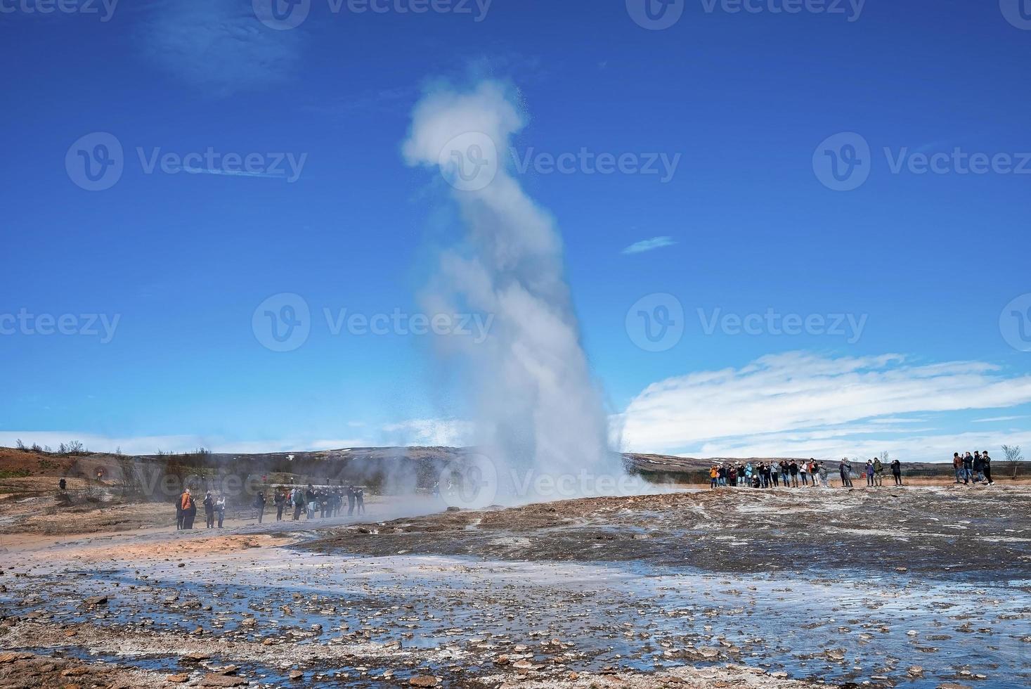 Tourists enjoying view of Strokkur geyser eruption in valley against blue sky photo