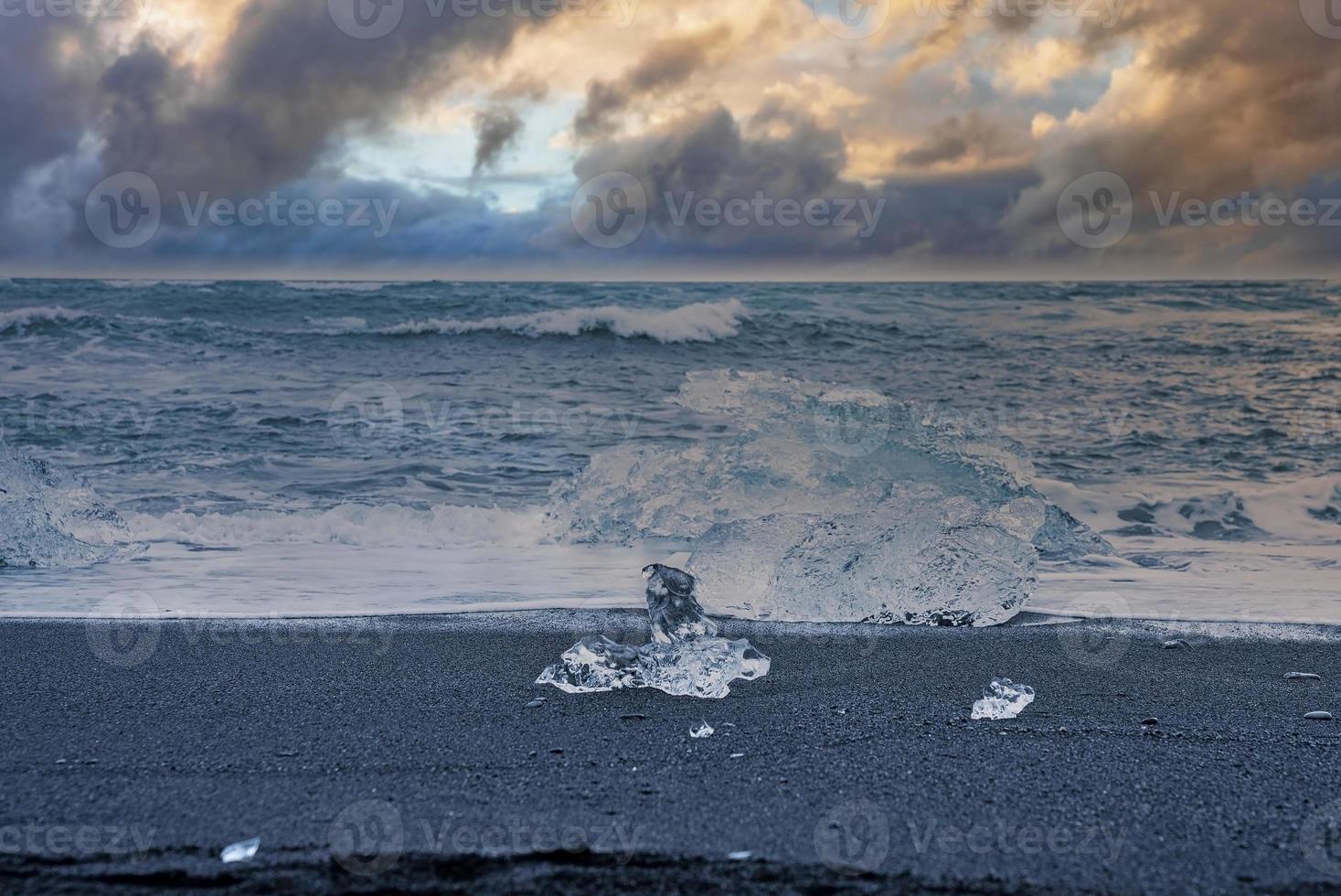 Iceberg chunk on black sand against waves rushing at Diamond beach during sunset photo