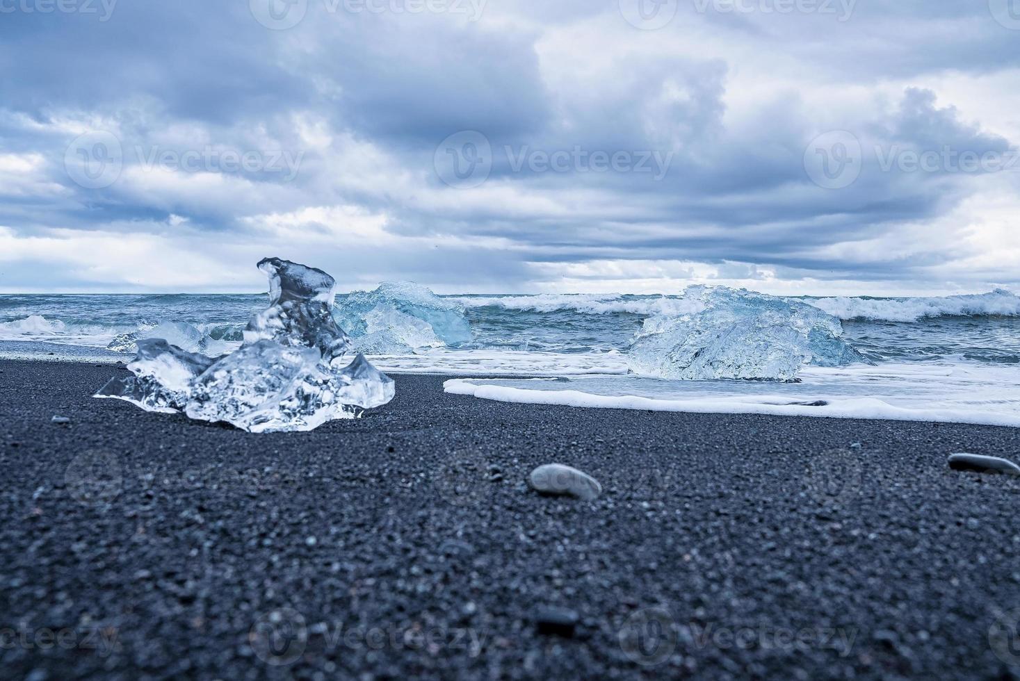 Iceberg chunks on black sand beach with waves rushing at shore against sky photo