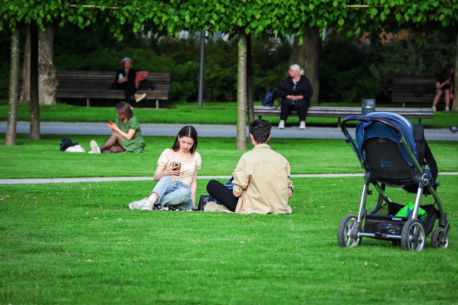 Vilnius, Lithuania. June 06 2022 - Young family couple sitting on green grass lawn woman looking at smart phone with child stroller standing on distance photo