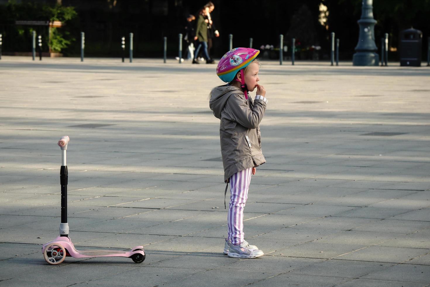 Vilnius, Lithuania. June 06 2022 - Small girl in pink helmet with pink scooter standing on city square thinking where to ride photo