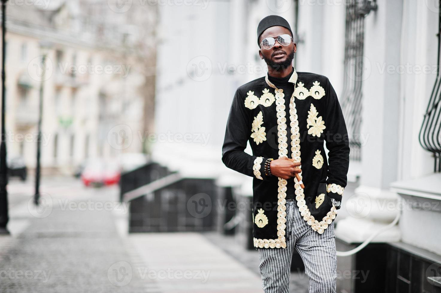 Mega stylish african man in traditional jacket pose. Fashionable black guy in hat and sunglasses with cigar in hand. photo