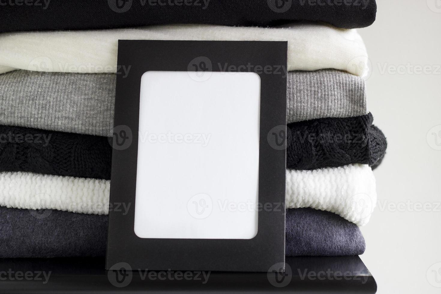 Close-up blank frame and stack of folded clothes on white background wall. Black, grey and white textiles. Copy space for text. Monochrome concept photo