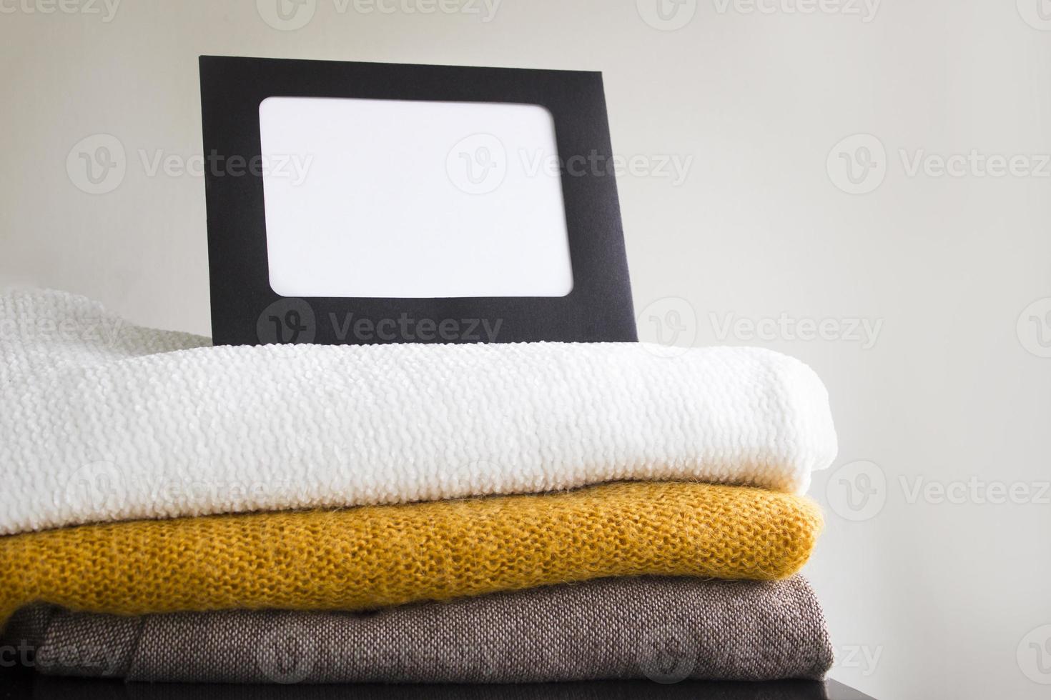 Close-up blank frame and stack of folded clothes on white background wall. Multicolored knitted textiles. Concept with copy space for text photo