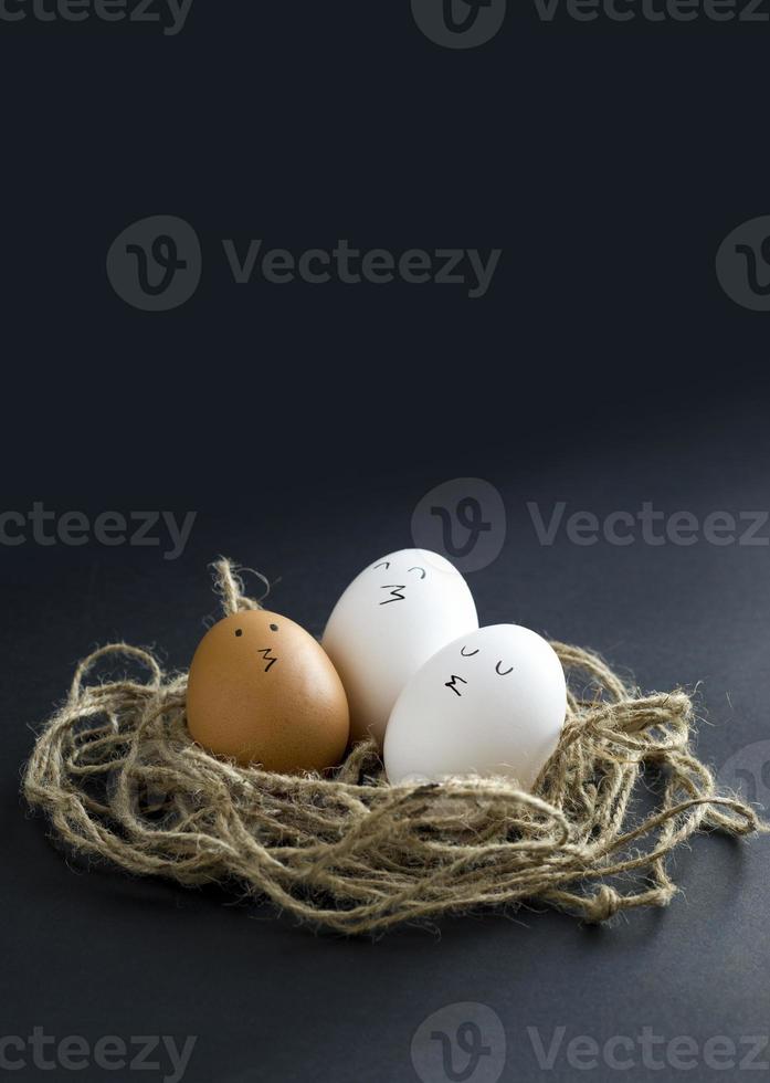 Nest with fresh chicken egg with drawn faces on black paper background. Concept for Easter with copy space. Creative layout made of white and brown eggs photo