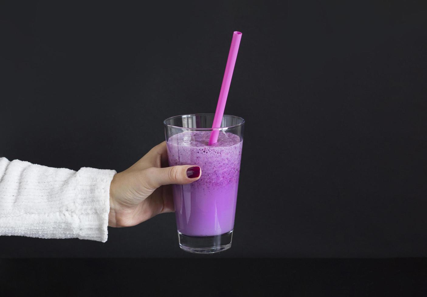 Female hand holds glass of berry smoothie with straw on black background. Pink milkshake with copy space. Healthy drink. Blueberry cocktail photo