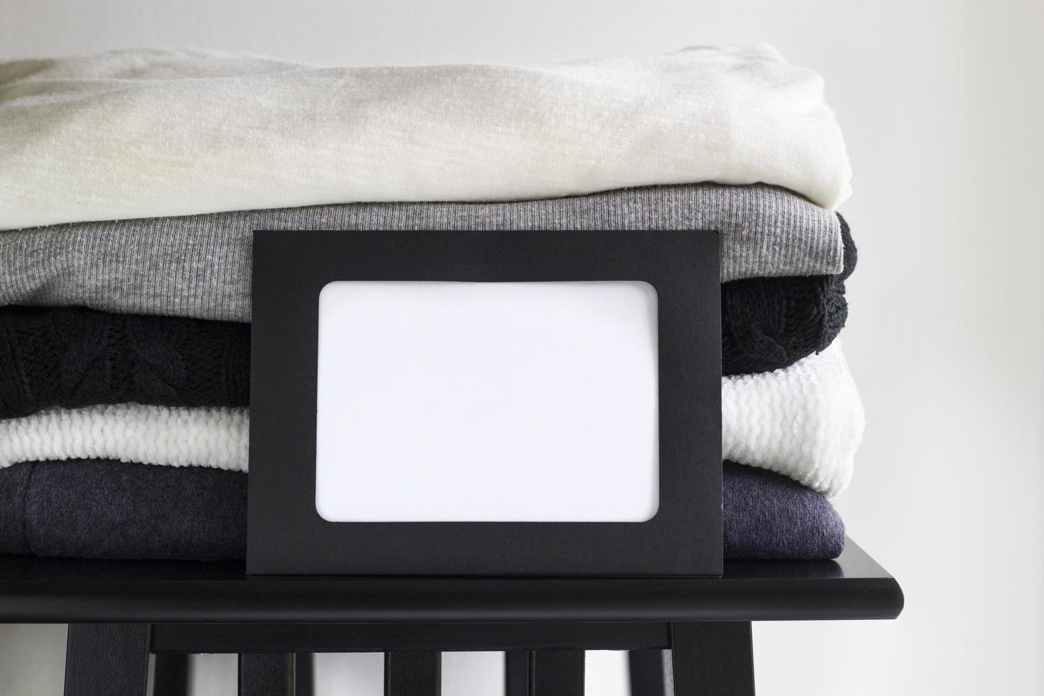 Close-up blank frame and stack of folded clothes on white background wall. Black, grey and white textiles. Copy space for text. Monochrome concept photo