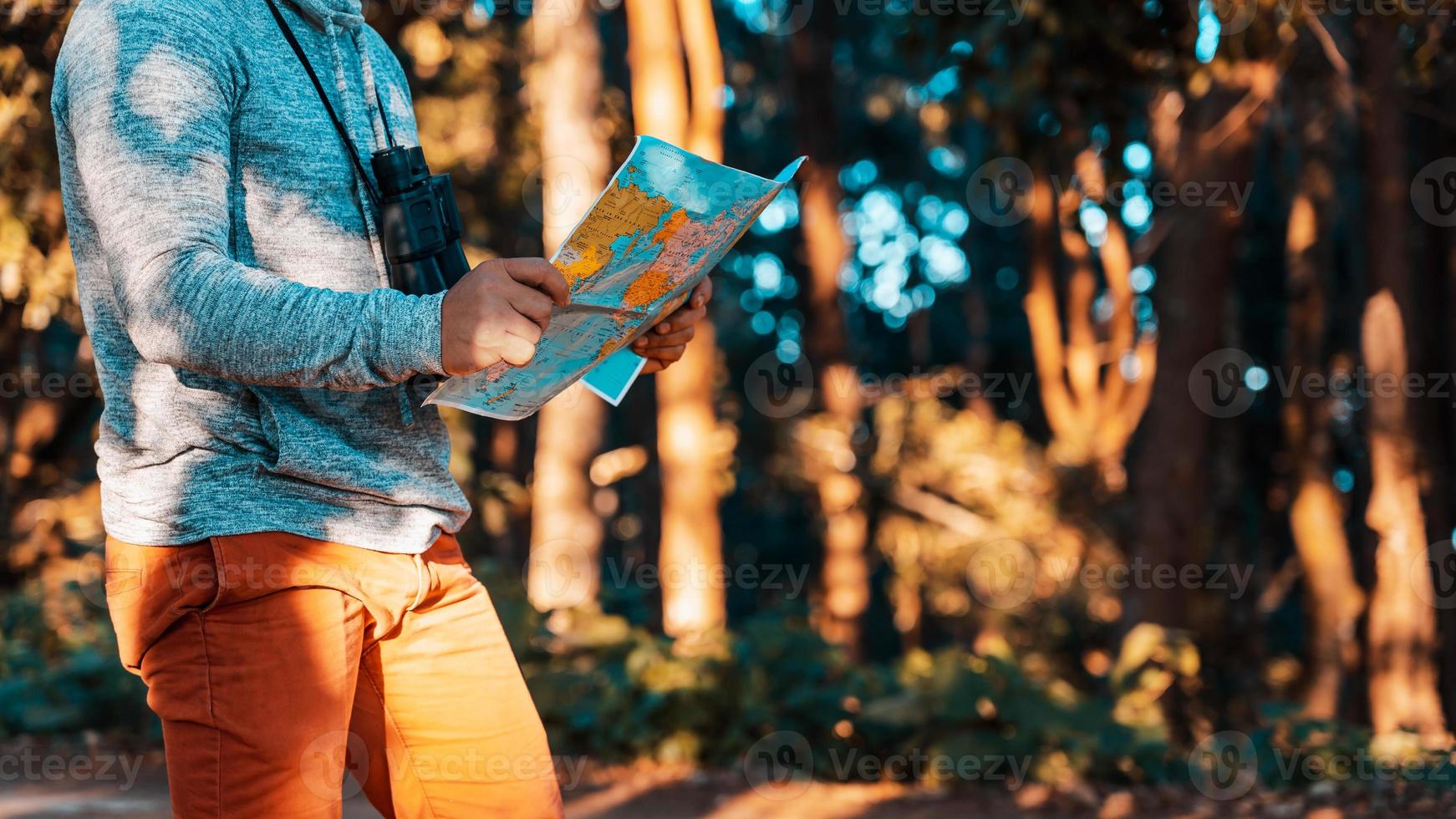 Traveler hipster man is in forest with binoculars and searching location in the map amongtrees big trees. photo