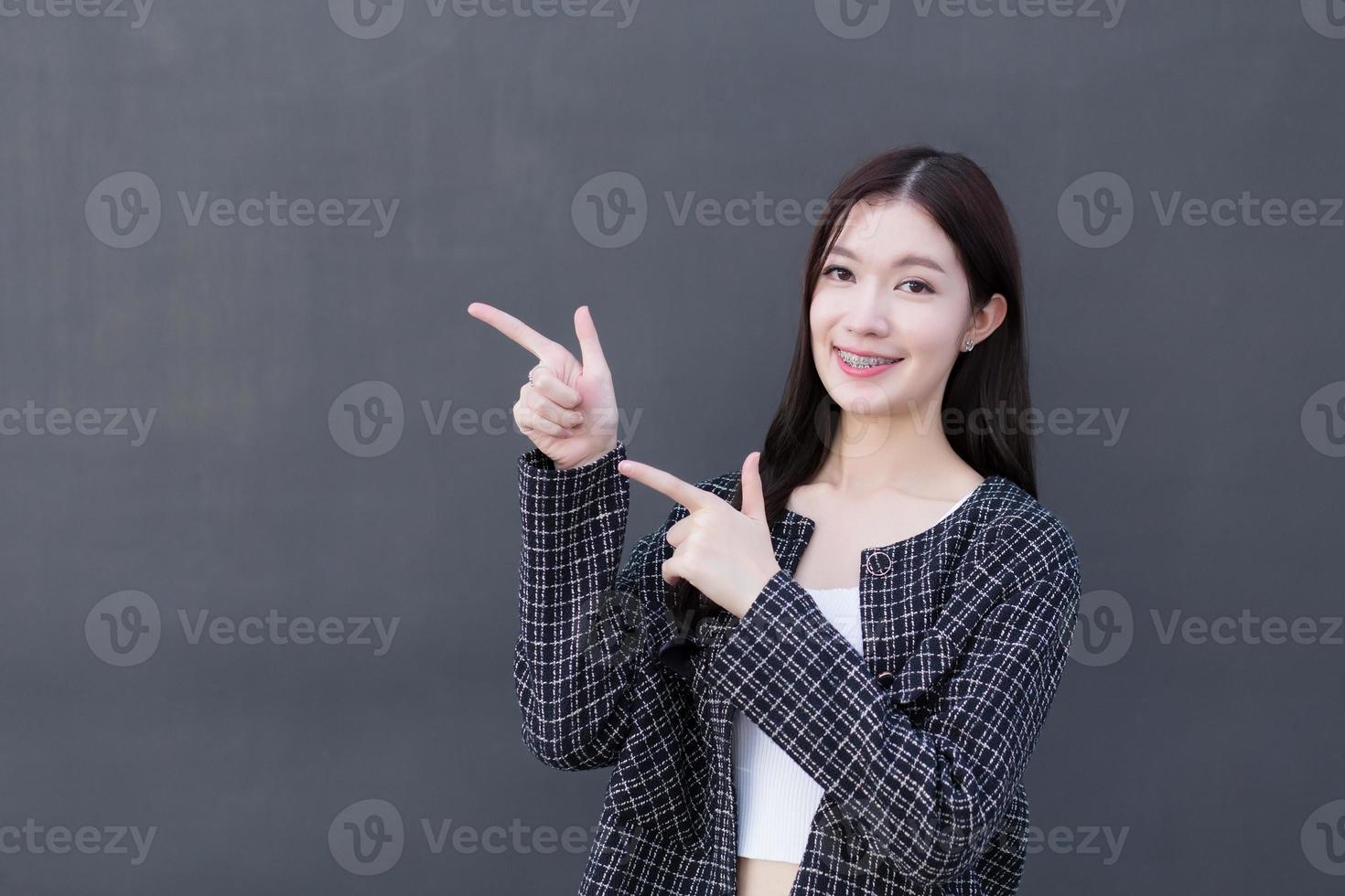 Asian working woman who wears black suit with braces on teeth is pointing hand to present something on the dark gray wall. photo