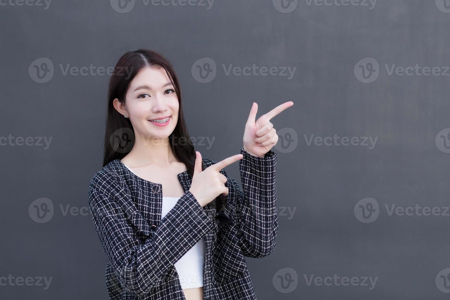 Asian working woman who wears black suit with braces on teeth is pointing hand to present something on the dark gray wall. photo