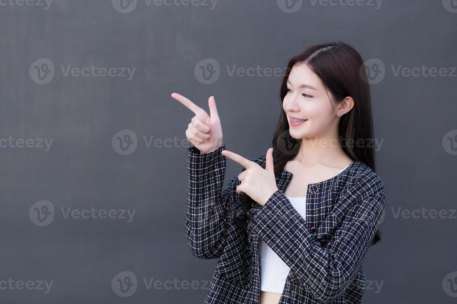 Asian professional working woman who wears black suit with braces on teeth is pointing hand to present something on the dark gray wall. photo