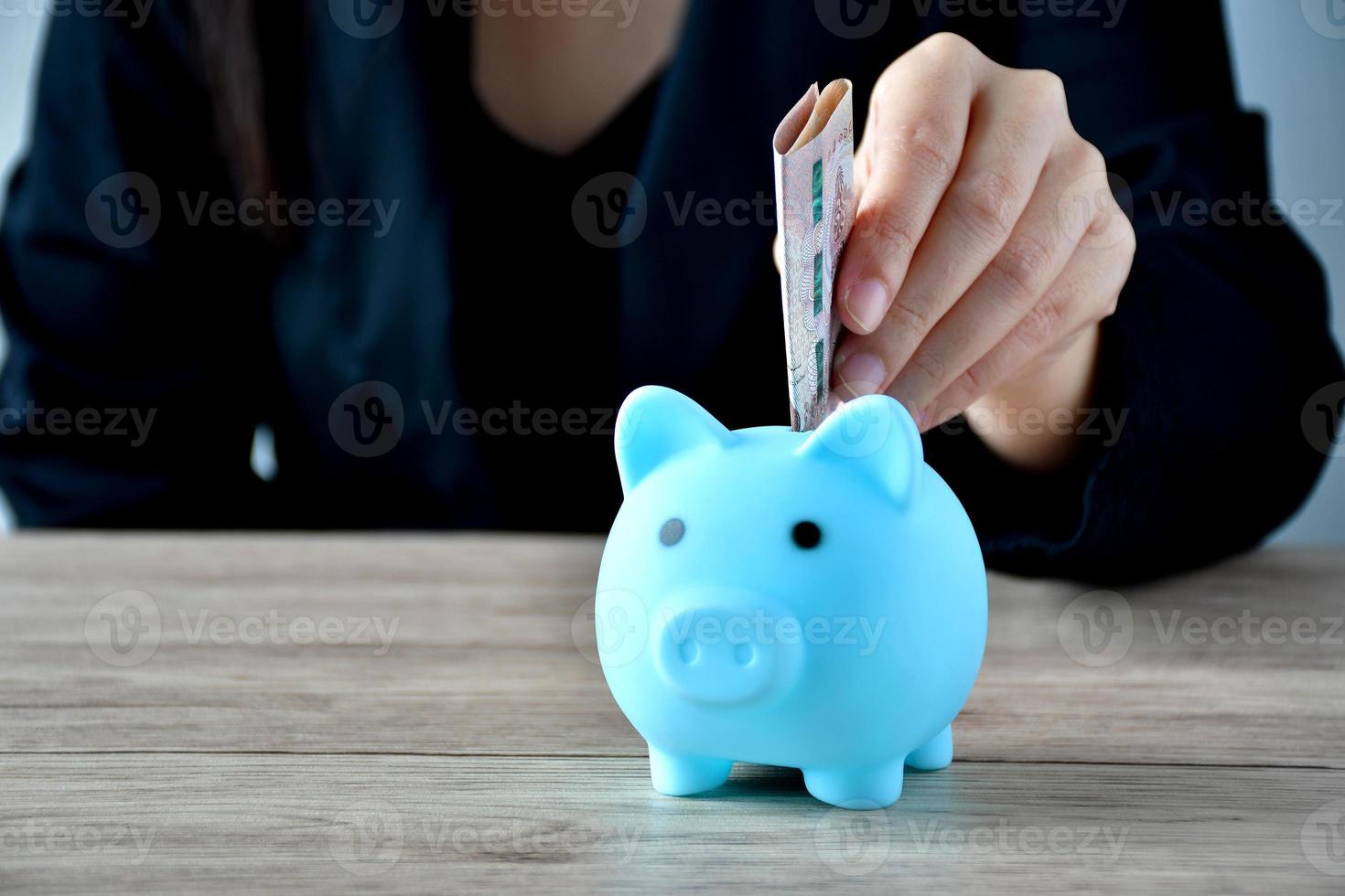 A woman puts a banknote in a blue piggy bank on a wooden table to save money on the tablet. photo