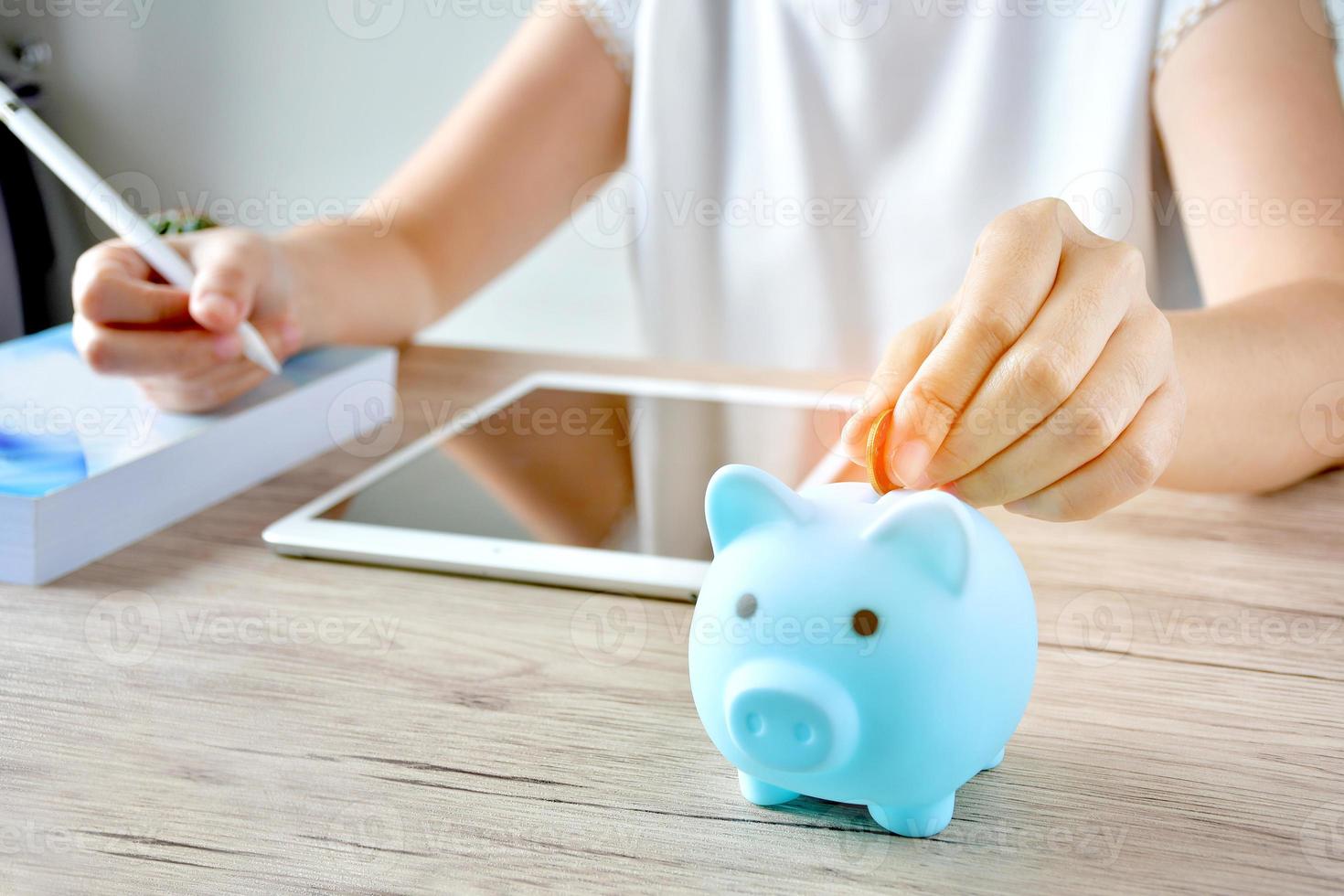 A woman puts a coin in a blue piggy bank on a wooden table to save money And record household income on the tablet. photo