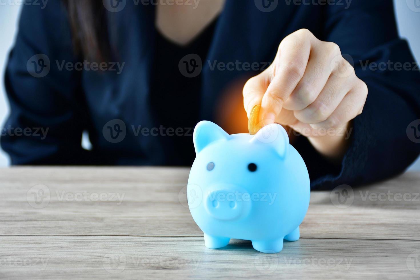 A woman puts a coin in a blue piggy bank on a wooden table to save money And record household income on the tablet. photo