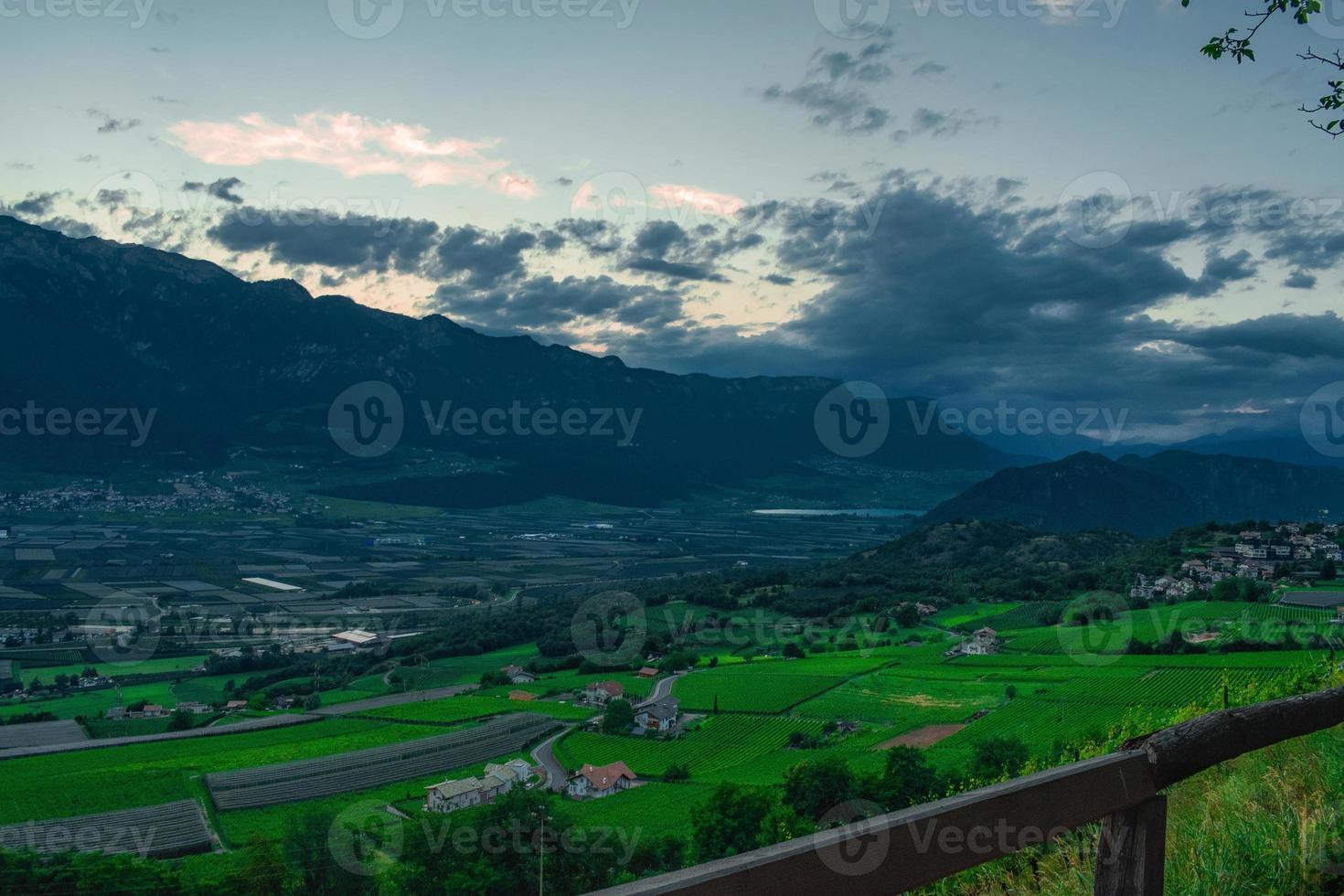 Mountain landscape imaged during sunset under a dramatic sky, dark clouds over mountain with a lake and a rural village in background photo