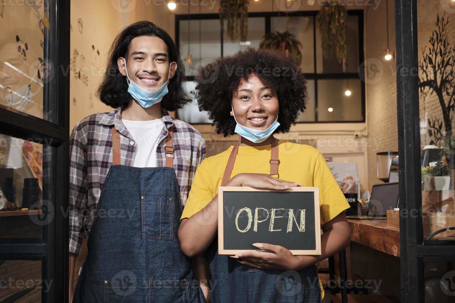 Two young startup barista partners stand at casual cafe door, write and show open sign before taking off face masks, happy and cheerful smiles with coffee shop service jobs after COVID-19 lifestyle. photo