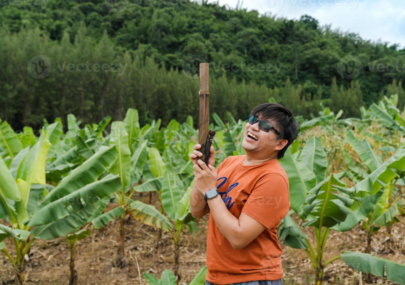 Asian man laughing with happiness holding bamboo bong in banana trees farm on green hill, bong use for marijuana or nicotinic smoking photo
