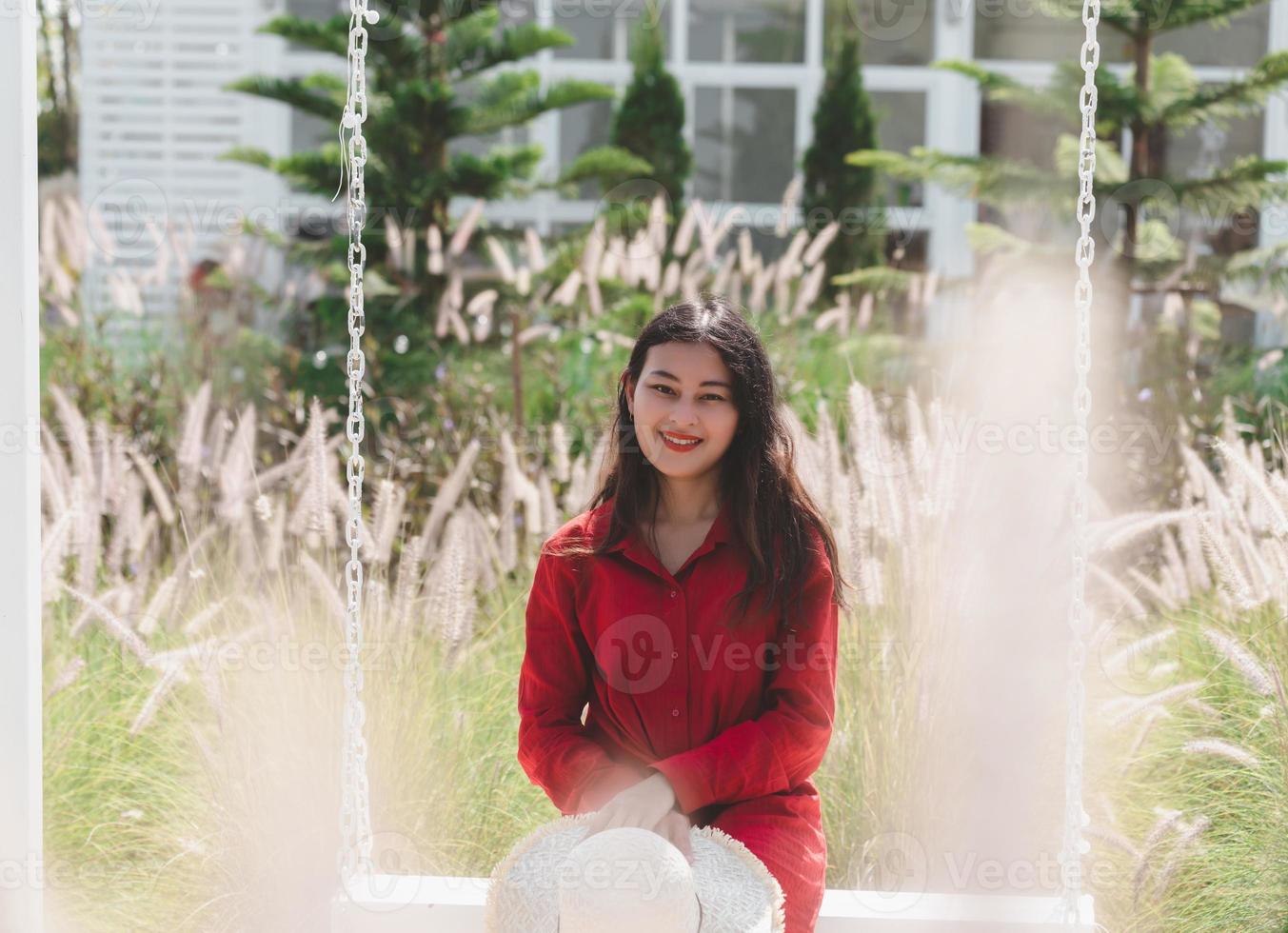 attractive Asian woman in red dress relax sit on swing in white glass flower garden photo