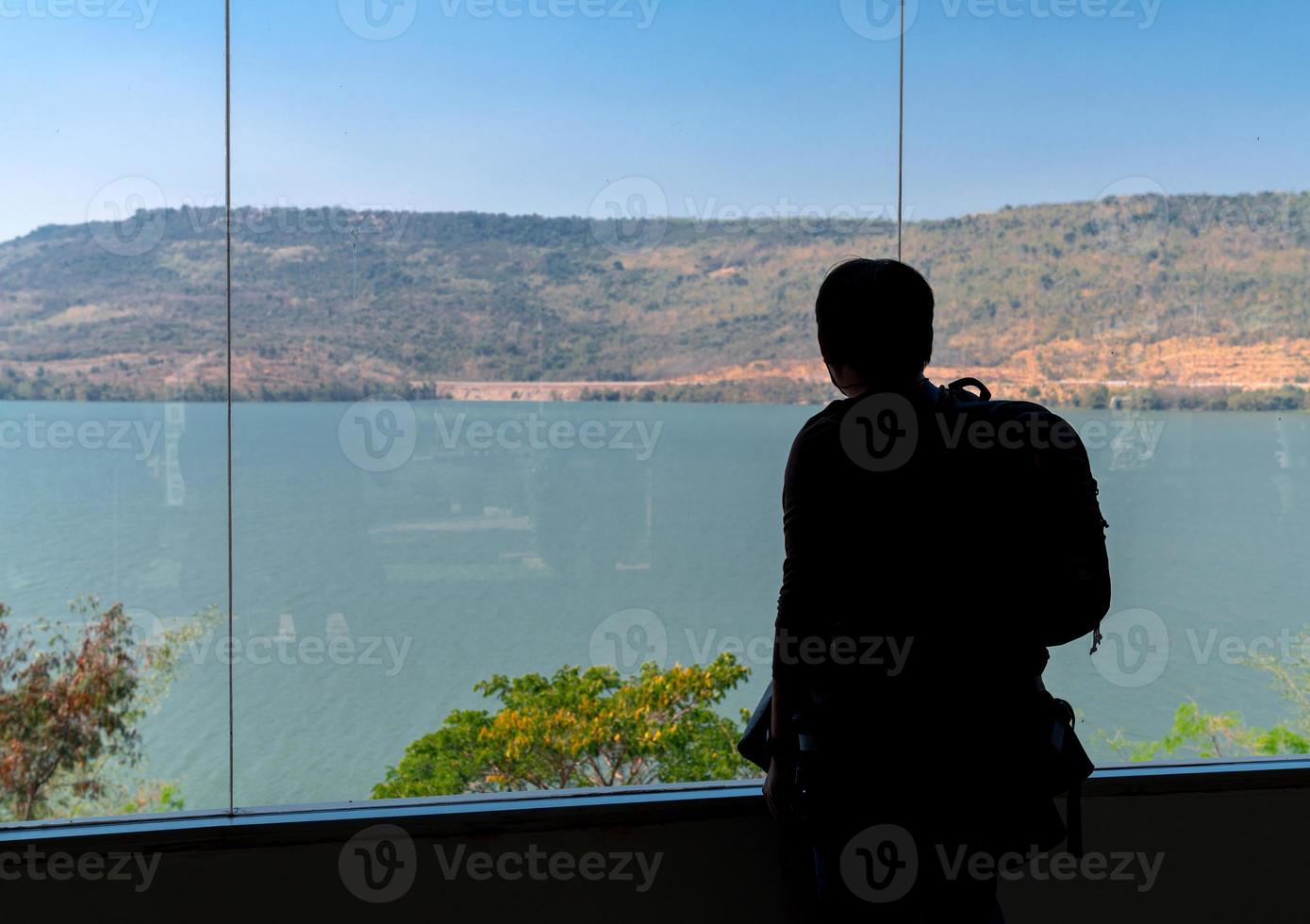 silueta trasera de un turista de mochila parado y mirando malditamente y vistas a la montaña en una ventana transparente desde la habitación interior foto