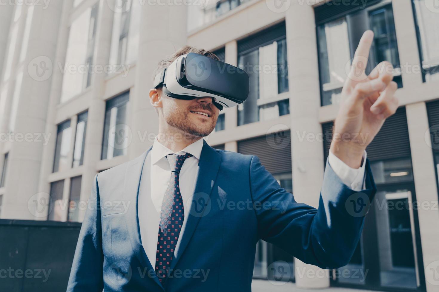 Cheerful businessman trying to touch something in virtual reality, standing near office building photo