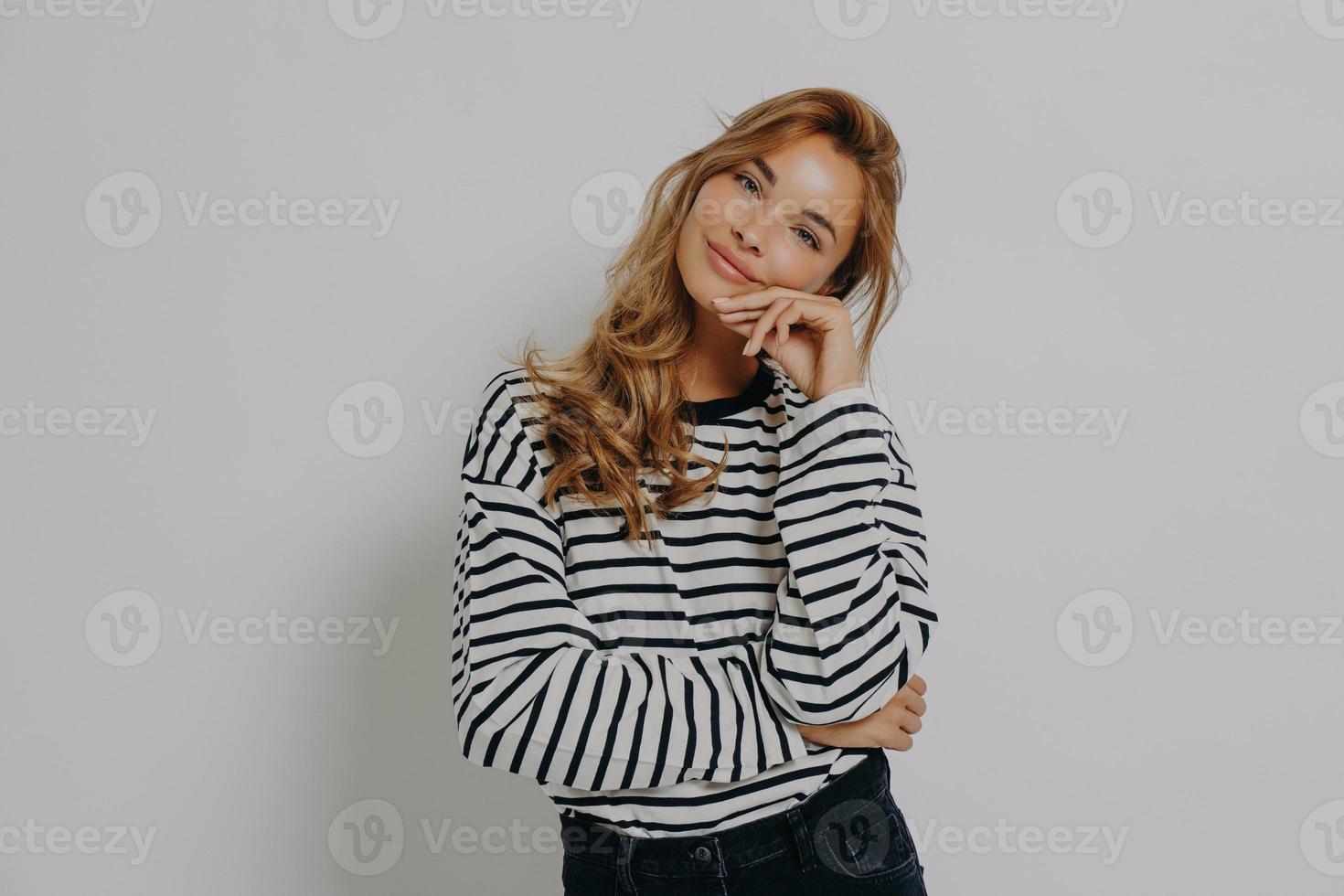 Studio shot of satisfied young European woman keeps hand on face tilts head looks gently at camera photo
