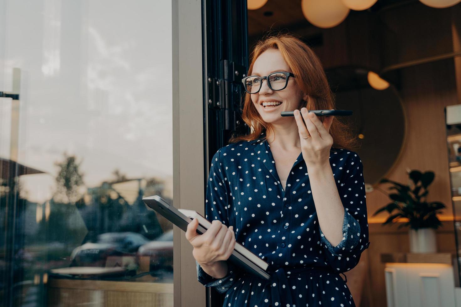 Happy ginger business woman freelancer with laptop holds mobile phone and talks on speakerphone photo