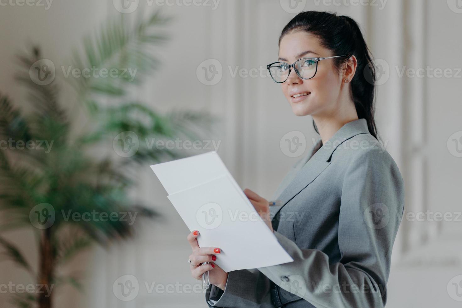 Successful brunette businesswoman stands thoughtful with paper documents, busy with making startup, analyzes business information, writes down notes, looks somewhere into distance, dressed formally photo