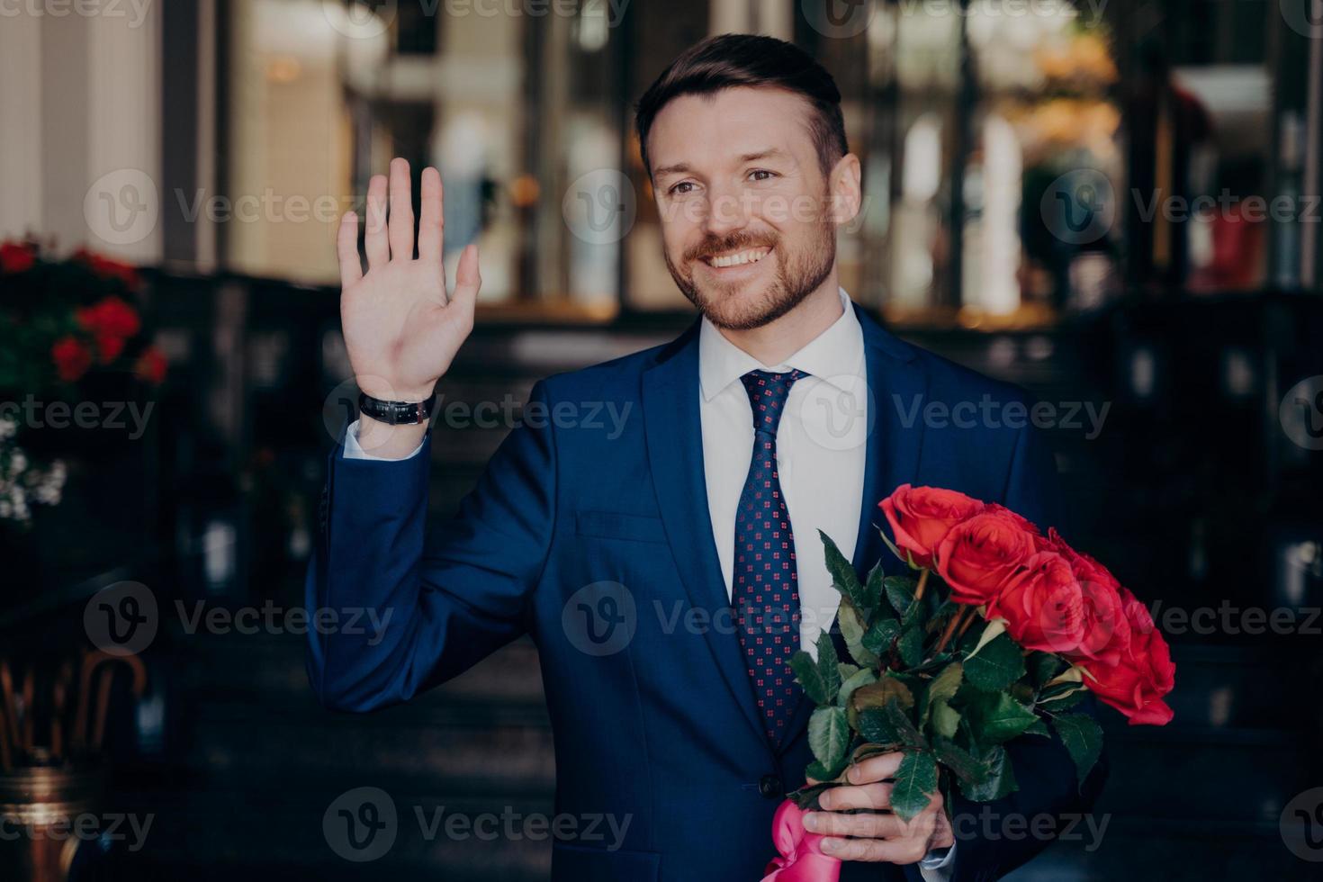 joven apuesto con barba ordenada en elegante esmoquin con rosas rojas en la mano foto
