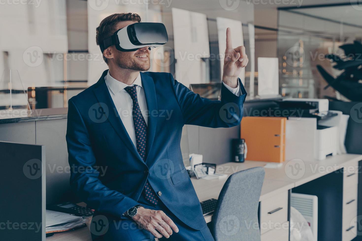 Enjoyed young businessman pointing with forefinger up in air with virtual reality glasses on head photo