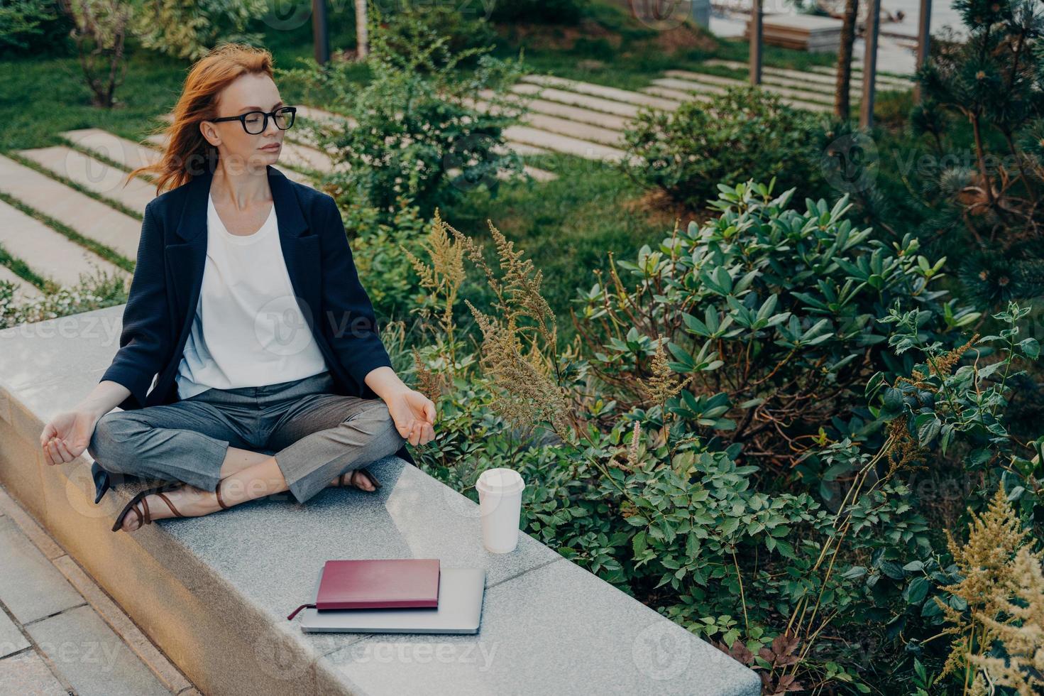 Relaxed ginger woman meditates in park has peaceful meditation enjoys nature photo