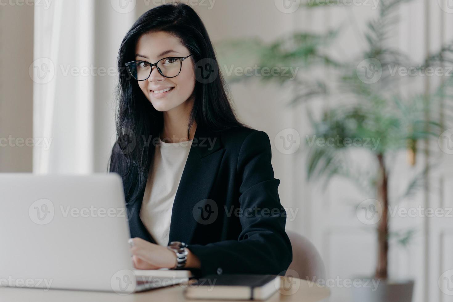 Smiling well dressed young woman secretary with dark hair, works on laptop computer at desktop, makes project or research work. Sales manager in modern office, cozy workplace. Remote job concept photo