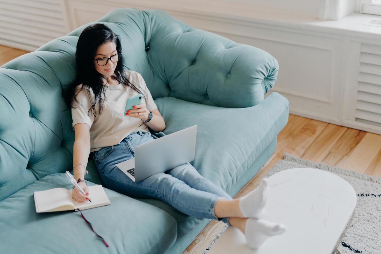 People, distance learning, technologies, freelance work concept. Serious brunette woman writes information in diary, holds mobile phone, concentrated on remote job, works with laptop computer photo
