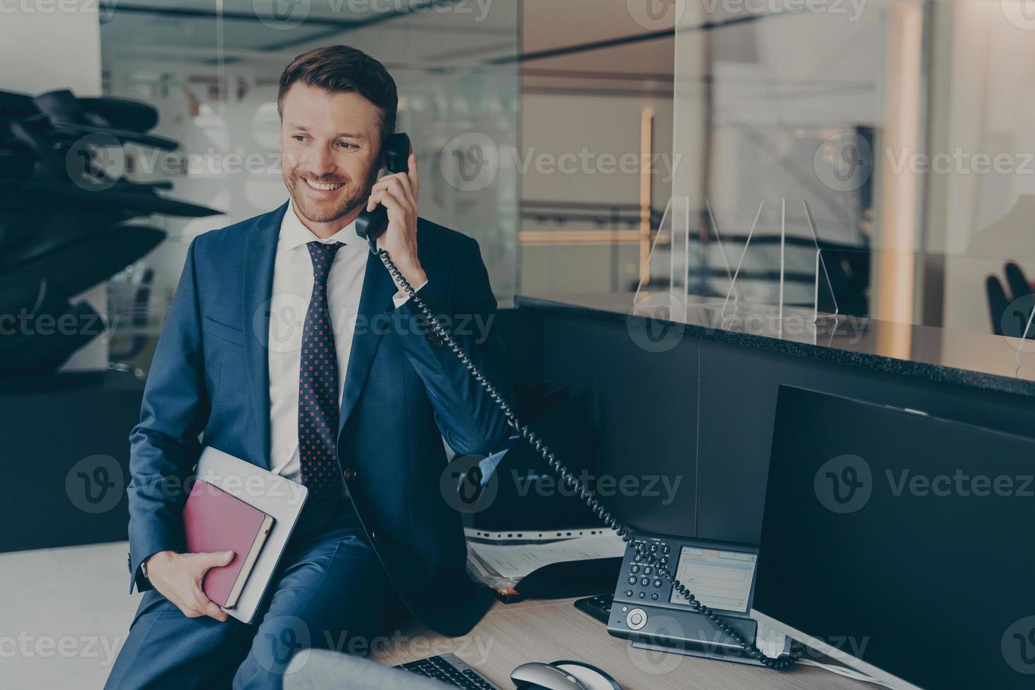 Businessman in formal wear, speaking on his office desk phone while sitting on top of table photo