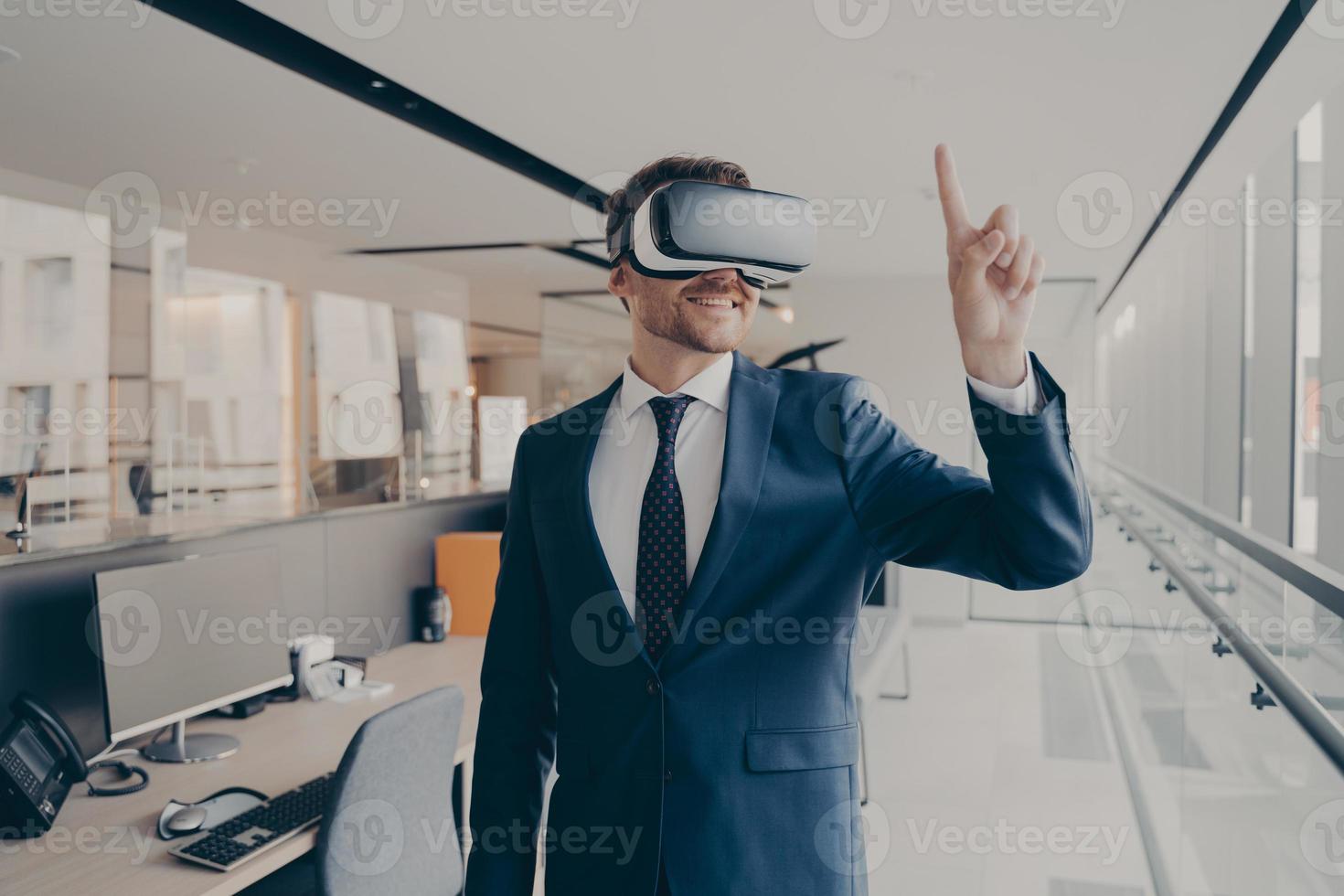 Happy man office worker wearing vr goggles, pointing with finger at objects in digital world photo