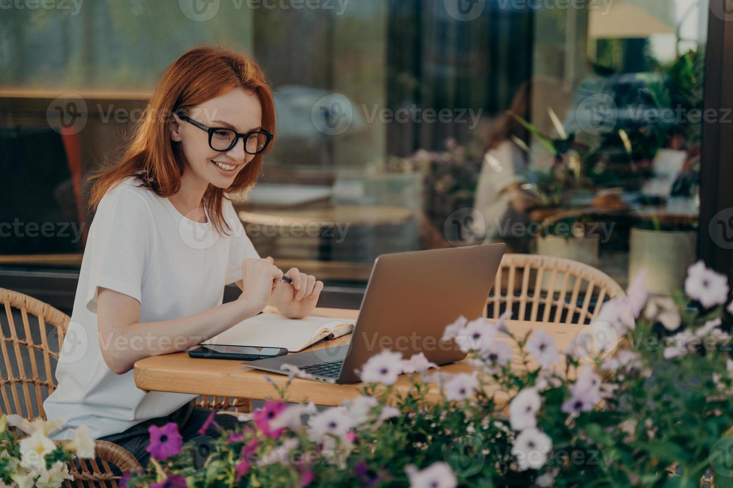 Redhead skillful woman student writes in notebook sits in front of opened laptop computer photo