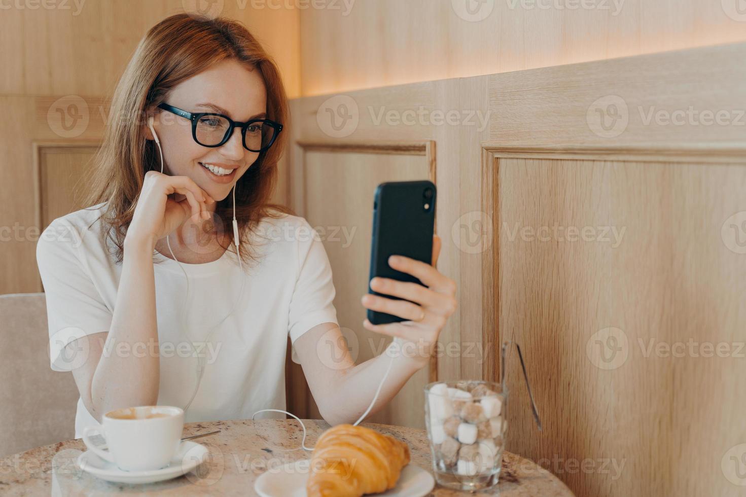 toma interior de una joven pelirroja alegre que usa teléfonos móviles y auriculares para conversar en línea foto