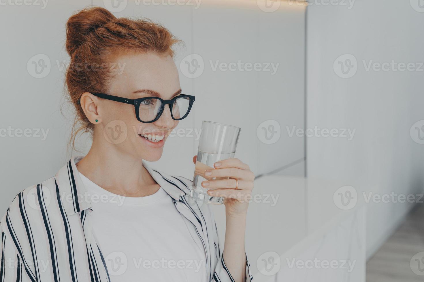 una joven pelirroja sonriente sostiene un vaso de agua bebe agua para refrescarse el cuerpo foto
