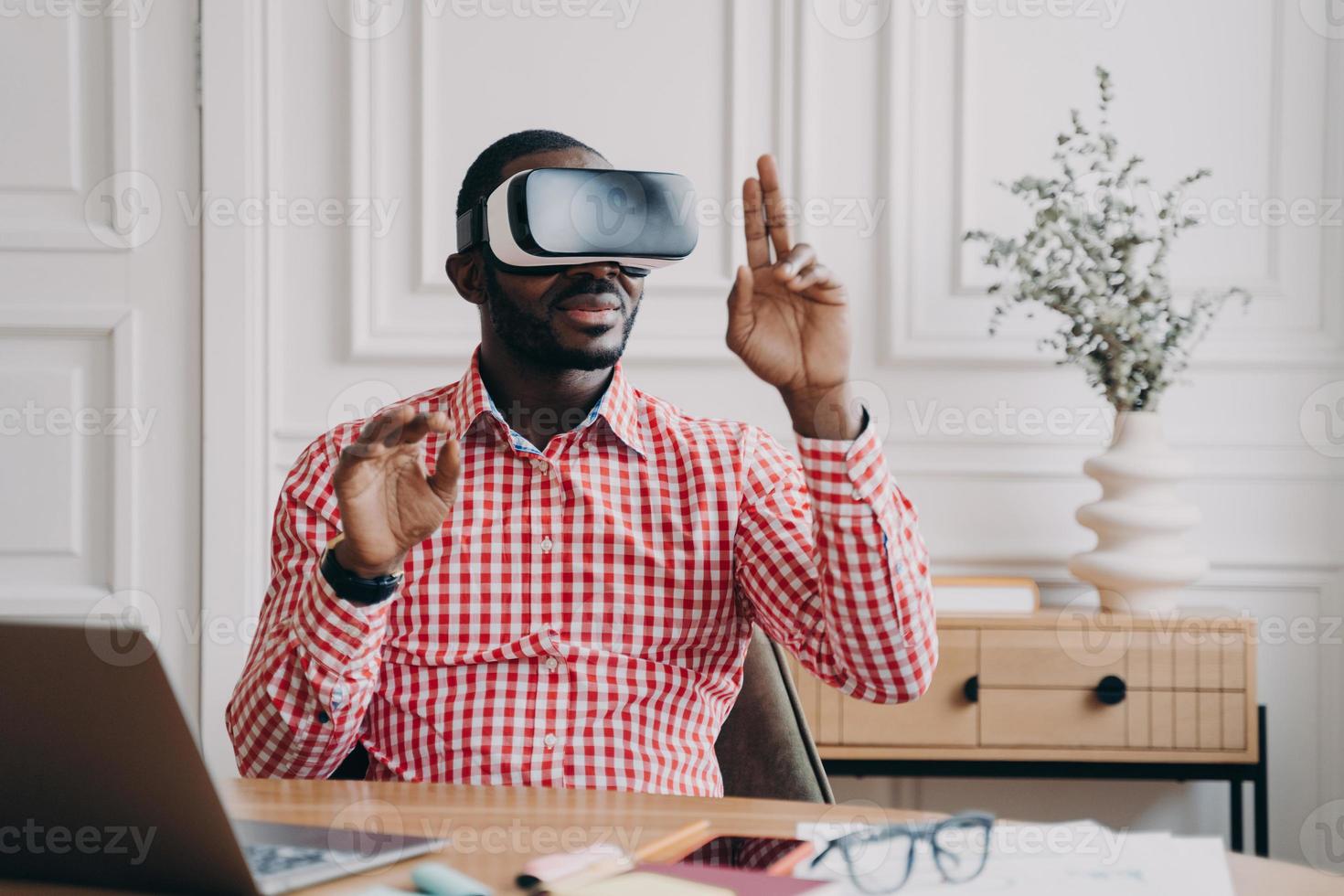 Focused Aframerican man in VR glasses enjoying augmented reality while sitting at workplace photo