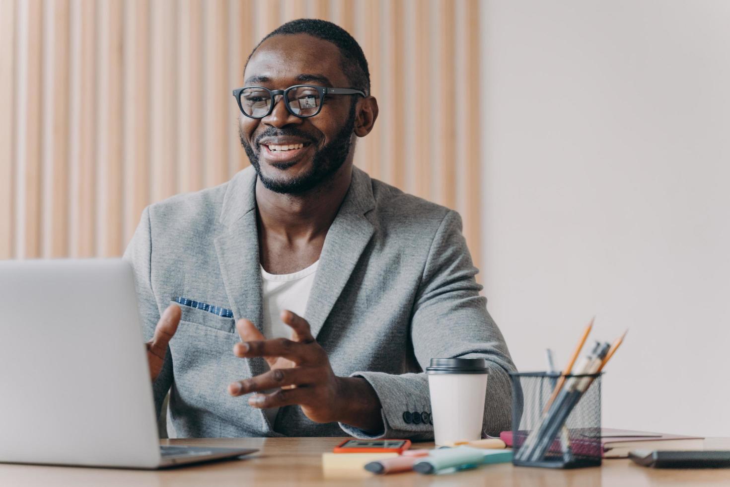 un alegre hombre de negocios afroamericano con gafas y un elegante blazer haciendo videollamadas en una laptop foto