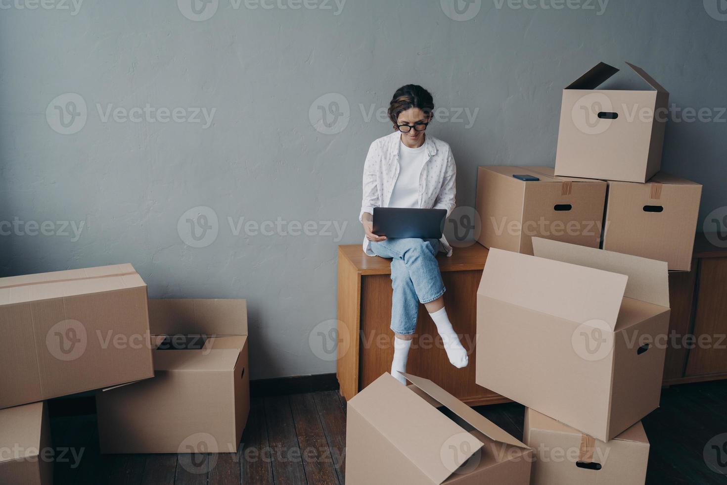 Spanish business lady rents apartment. Happy girl is sitting among boxes and working on laptop. photo