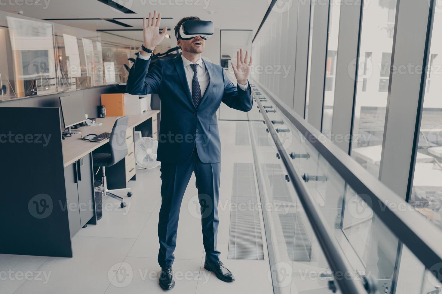 Young amazed businessman in VR headset trying to touch something with hands while standing in modern office photo