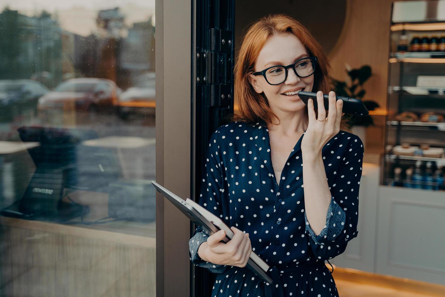 Female freelancer stands outdoors with mobile phone and uses voice recognition system on smartphone photo